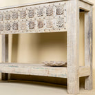 Close up side angle of a  handcarved console table made from reclaimed teak wood, featuring a detailed front panel with geometric floral motifs and a distressed whitewashed finish, complemented by a lower shelf for added storage or display.
