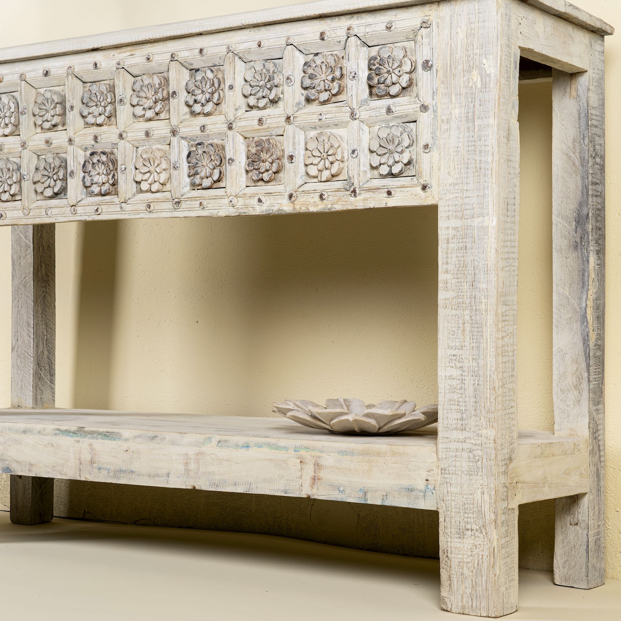 Close up side angle of a  handcarved console table made from reclaimed teak wood, featuring a detailed front panel with geometric floral motifs and a distressed whitewashed finish, complemented by a lower shelf for added storage or display.
