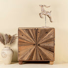 Front view image of a artistic wooden cabinet in rustic style, made from reclaimed wood with a striking sunburst pattern on the double doors, featuring subtle color variations and round metal ring pulls, elevated on rounded wooden feet.
