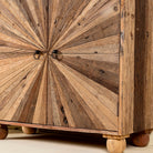 Close up leg image of a artistic wooden cabinet in rustic style, made from reclaimed wood with a striking sunburst pattern on the double doors, featuring subtle color variations and round metal ring pulls, elevated on rounded wooden feet.
