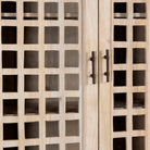 Close up image of a sandblasted mango wood showcase with two glass-panelled grid doors and four interior compartments, finished in a light sun-washed tone, ideal as living room furniture.