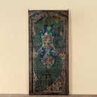 A tall, narrow hand-painted wooden door panel featuring two peacocks with elaborate teal, blue, and gold feather patterns on a distressed brown background, set against a plain beige wall.