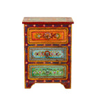 White background image of a hand-painted reclaimed wood nightstand with multiple drawers, featuring vibrant colours and floral patterns, placed next to a vase with dried plants.