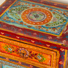 Close up image of a hand-painted reclaimed wood nightstand with multiple drawers, featuring vibrant colours and floral patterns, placed next to a vase with dried plants.