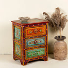 Side angle image of a hand-painted reclaimed wood nightstand with multiple drawers, featuring vibrant colours and floral patterns, placed next to a vase with dried plants.