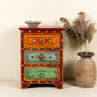 A hand-painted reclaimed wood nightstand with multiple drawers, featuring vibrant colours and floral patterns, placed next to a vase with dried plants.