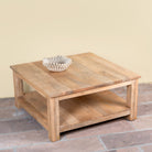 A square wooden coffee table with a natural light brown finish and an open lower shelf, styled with a small white ribbed bowl on top, set against a beige wall and brick floor.