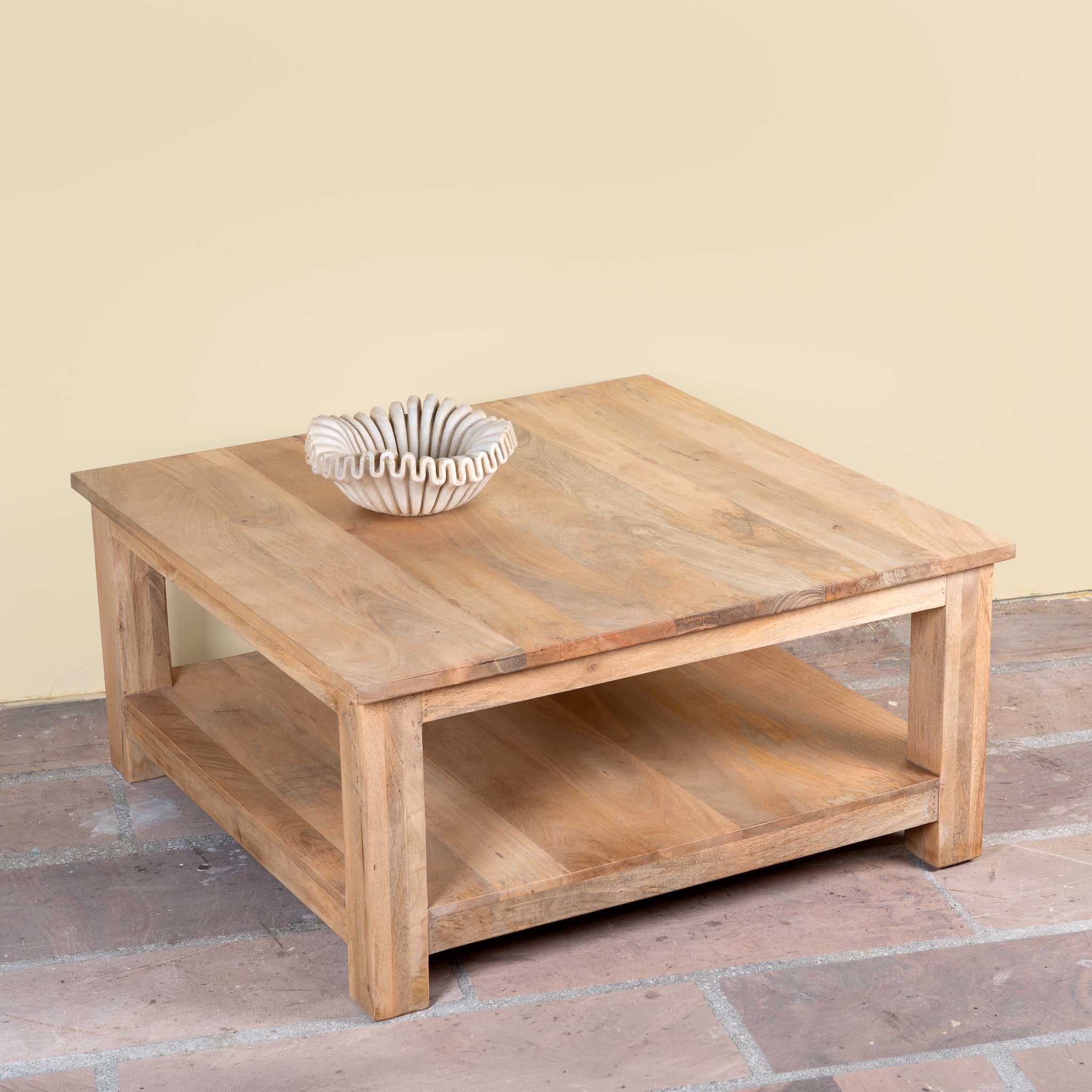 A square wooden coffee table with a natural light brown finish and an open lower shelf, styled with a small white ribbed bowl on top, set against a beige wall and brick floor.