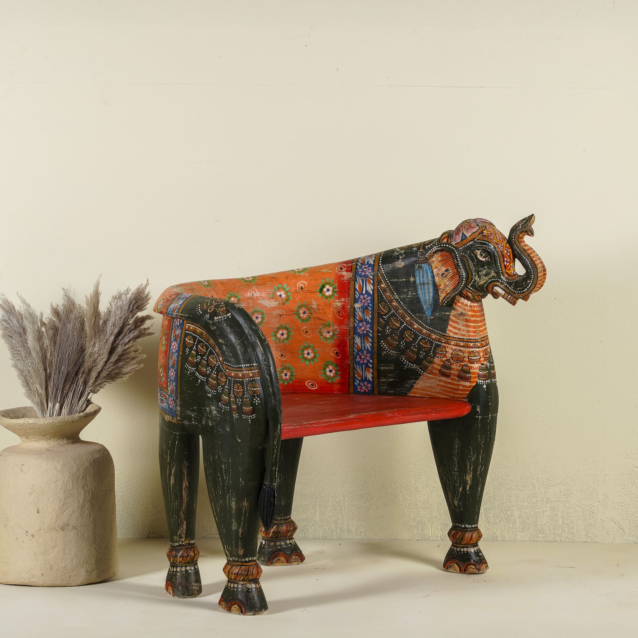 Side angle image of a elephant-shaped armchair crafted from reclaimed wood, featuring intricate hand-painted details in vibrant red, orange, green, and multi coloured floral patterns, with sculpted elephant head and legs for a whimsical, artistic touch.
