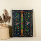 Hand-painted wall panel made from an old teak wood window frame, adorned with detailed floral motifs in shades of pink, red, green, and white against a deep blue background, framed by intricate carvings and colourful decorative accents.

