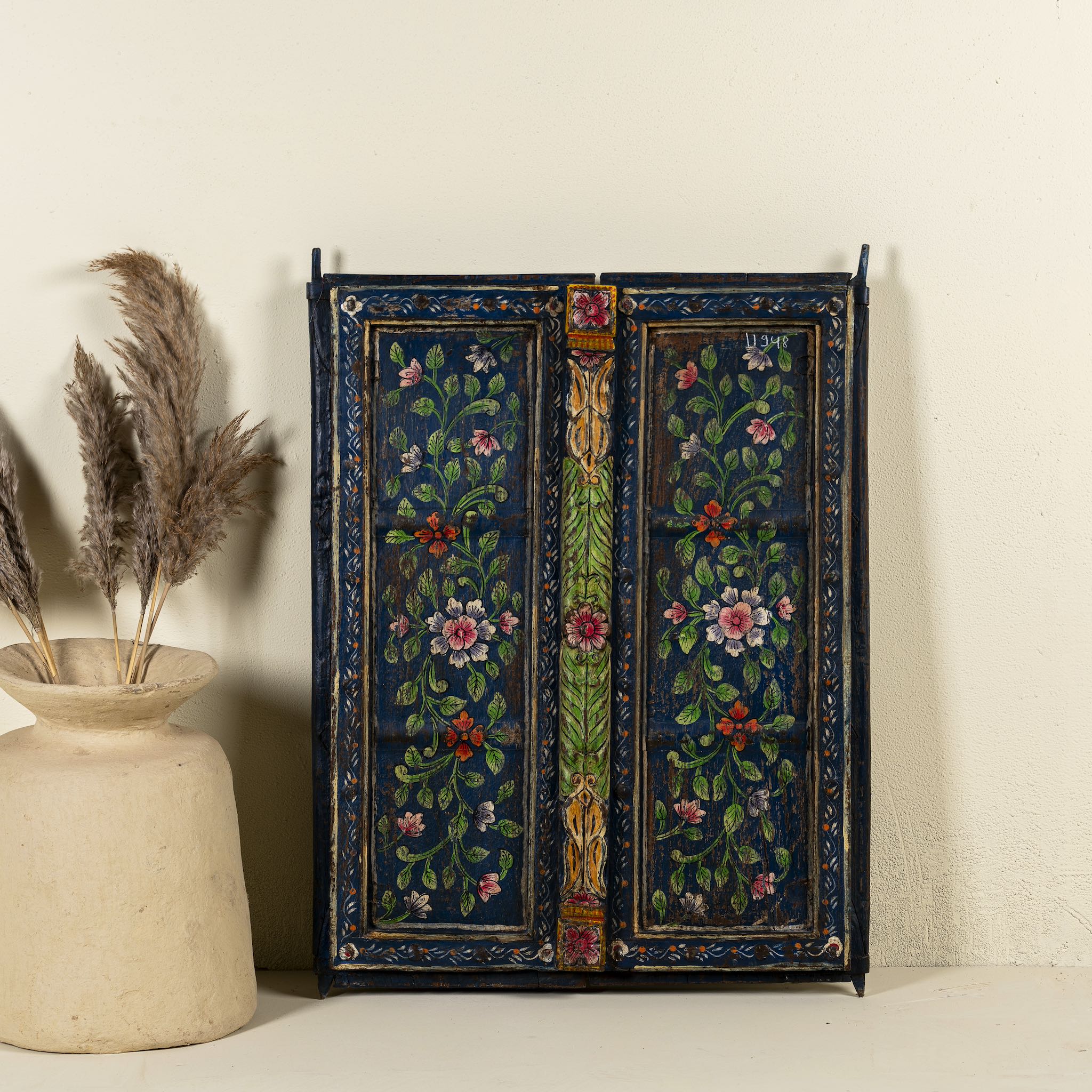 Hand-painted wall panel made from an old teak wood window frame, adorned with detailed floral motifs in shades of pink, red, green, and white against a deep blue background, framed by intricate carvings and colourful decorative accents.
