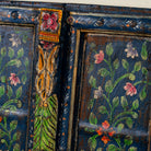 Close up image of a hand-painted wall panel made from an old teak wood window frame, adorned with detailed floral motifs in shades of pink, red, green, and white against a deep blue background, framed by intricate carvings and colourful decorative accents.
