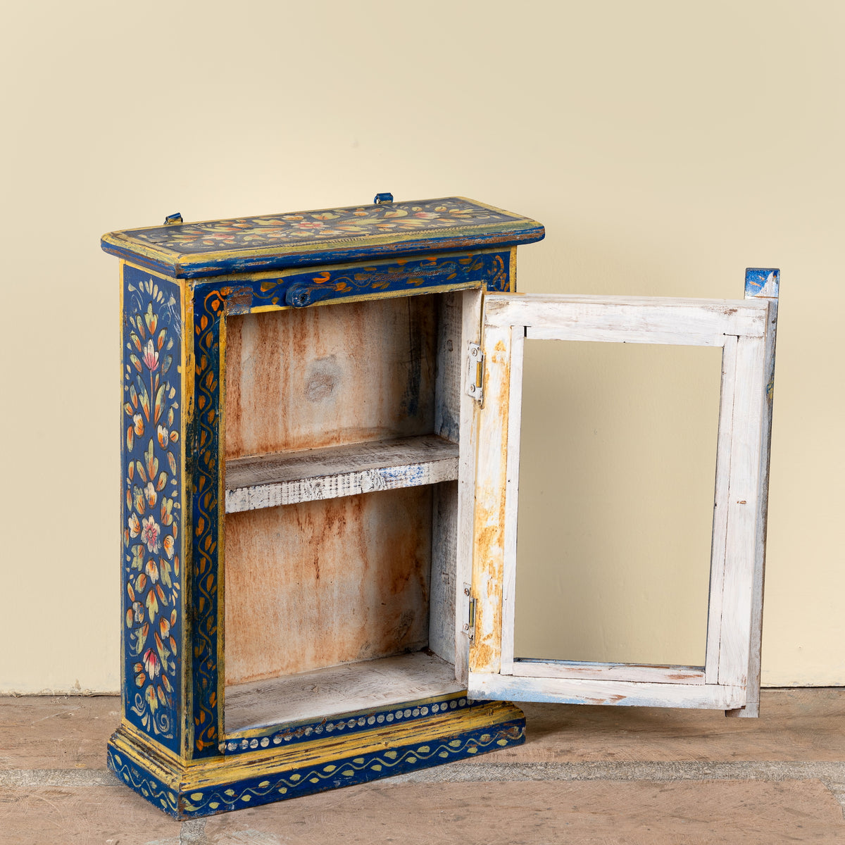 A small decorative cabinet with a distressed white door swung open, showcasing intricate blue, gold, and floral hand-painted details on the exterior, set against a plain beige wall.
