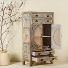 Open door image of a hand-painted cabinet made from mango wood, featuring rattan inlay doors framed by a carved circular motif, floral blue and green accents, and five drawers for additional storage.
