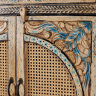 Close up image of a hand-painted cabinet made from mango wood, featuring rattan inlay doors framed by a carved circular motif, floral blue and green accents, and five drawers for additional storage.