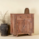 Side angle image of a acacia wood cabinet with hand-carved sunburst doors, grooved details, and one drawer, 90 × 40 × 90 cm.