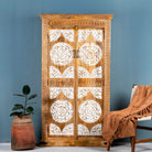 Hand-carved solid mango wood wardrobe with whitewashed floral carvings on the doors, a brown textile on the floor, and a plant pot beside it, against a blue wall.