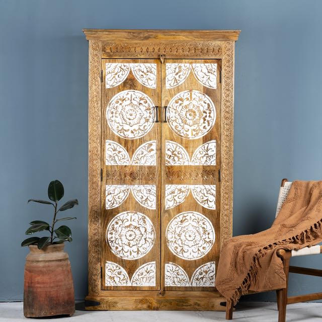 Hand-carved solid mango wood wardrobe with whitewashed floral carvings on the doors, a brown textile on the floor, and a plant pot beside it, against a blue wall.