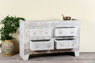 White distressed dresser with decorative carvings on a wooden floor with a plant and vase in the background.