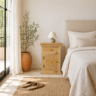 Lifestyle image of a hand-carved nightstand made of bleached mango wood with floral Mediterranean patterns and one drawer – Hriday bedside tables by Om Home & Garden