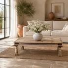 Lifestyle image of a rustic coffee table made from old reclaimed teak wood, featuring a weathered finish, turned legs, decorative carved apron, and visible metal rivets, styled with earthy decor in a minimal interior setting.
