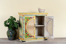 Side angle image with open door of a hand-painted, hand-carved cupboard made of mango wood with floral design and rattan inserts.