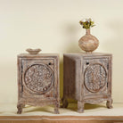 Pair of hand-carved bedside tables made of mango wood, featuring intricate round floral motifs on the doors, crackled vintage finish, and elegant curved legs in a distressed antique design.