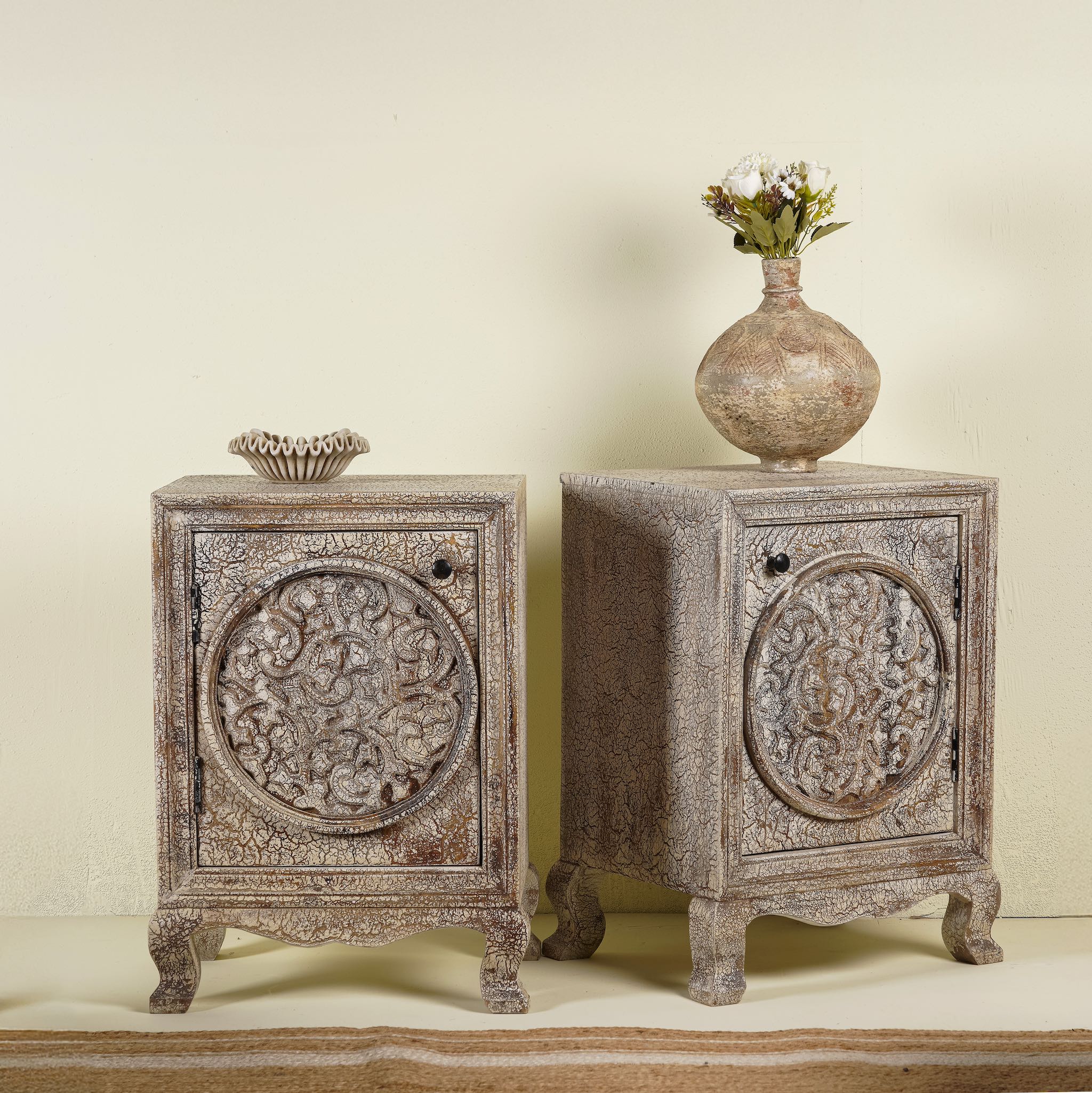 Pair of hand-carved bedside tables made of mango wood, featuring intricate round floral motifs on the doors, crackled vintage finish, and elegant curved legs in a distressed antique design.