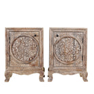 White background picture of a pair of hand-carved bedside tables made of mango wood, featuring intricate round floral motifs on the doors, crackled vintage finish, and elegant curved legs in a distressed antique design.
