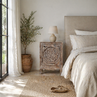 Lifestyle scene of a hand-carved bedside table made of mango wood, featuring intricate round floral motifs on the doors, crackled vintage finish, and elegant curved legs in a distressed antique design.