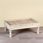 Side angle image of a Jayson reclaimed teak wood coffee table with glass top, removable legs, and hand carved antique Indian ceiling panel.