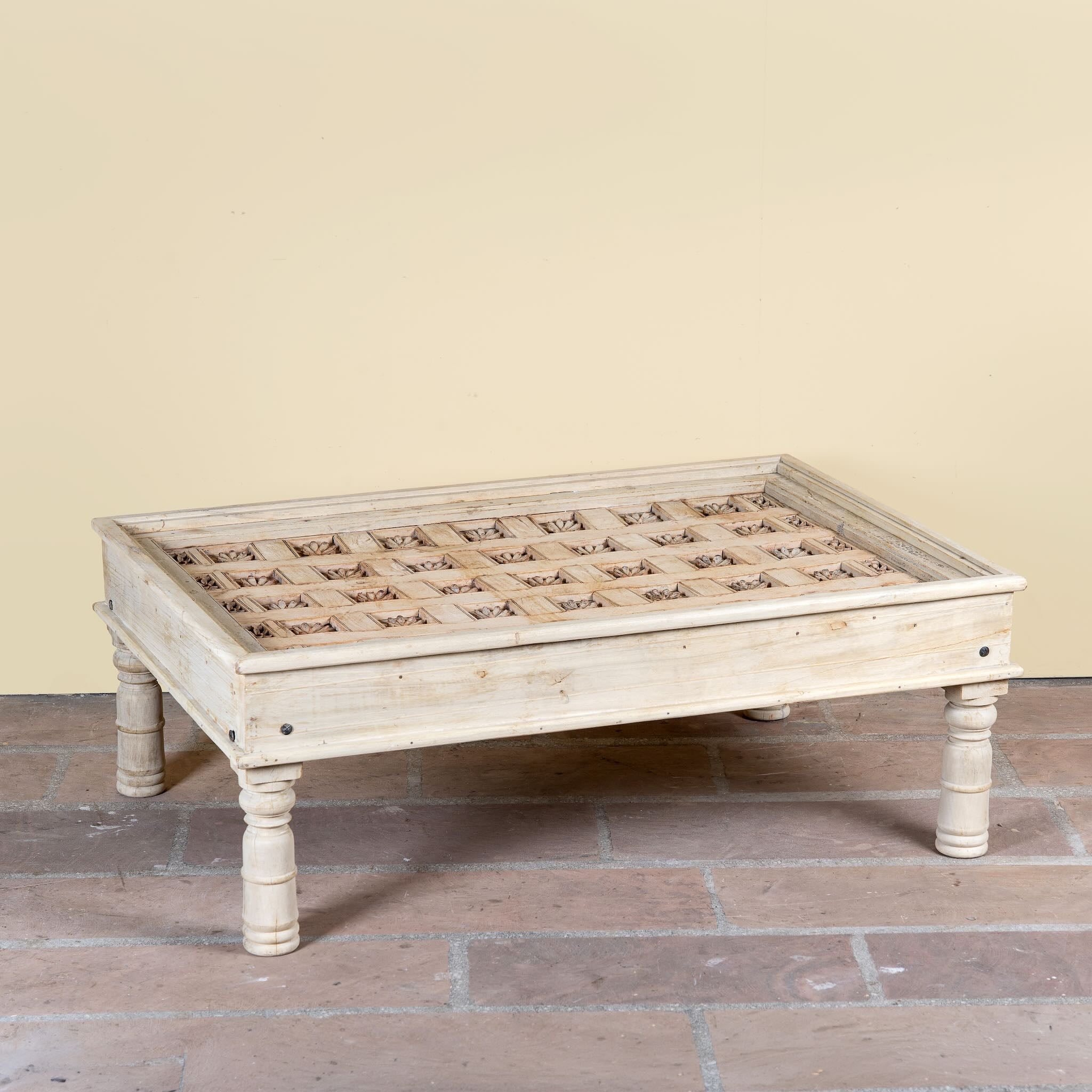 Side angle image of a Jayson reclaimed teak wood coffee table with glass top, removable legs, and hand carved antique Indian ceiling panel.