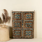 Antique wall panel made from an old teak wood window frame, featuring hand-painted blue floral motifs on a distressed natural wood background, with ornate yellow, white, and blue border accents and decorative metal ring pulls.
