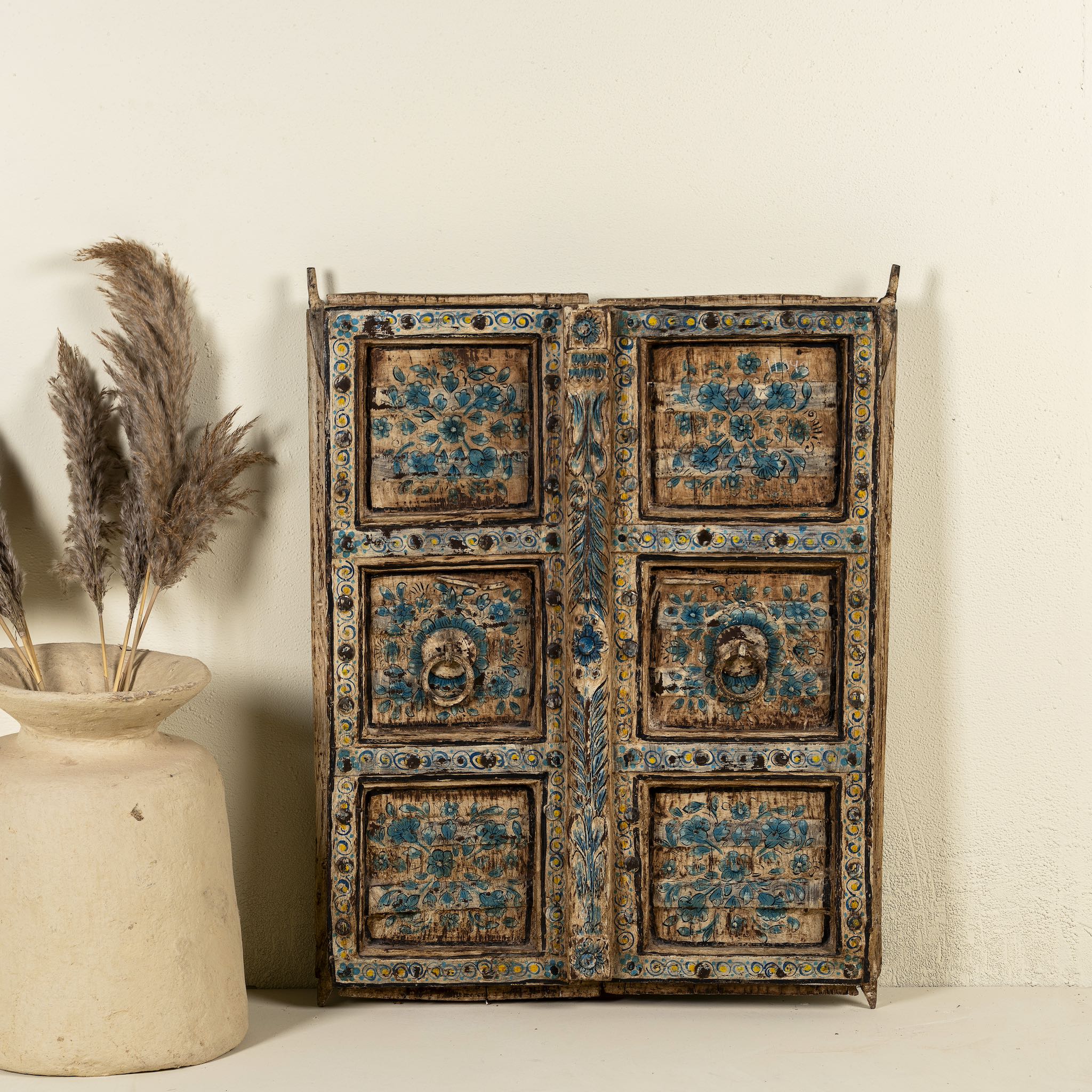 Antique wall panel made from an old teak wood window frame, featuring hand-painted blue floral motifs on a distressed natural wood background, with ornate yellow, white, and blue border accents and decorative metal ring pulls.
