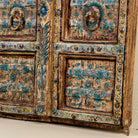 Close up image of a antique wall panel made from an old teak wood window frame, featuring hand-painted blue floral motifs on a distressed natural wood background, with ornate yellow, white, and blue border accents and decorative metal ring pulls.
