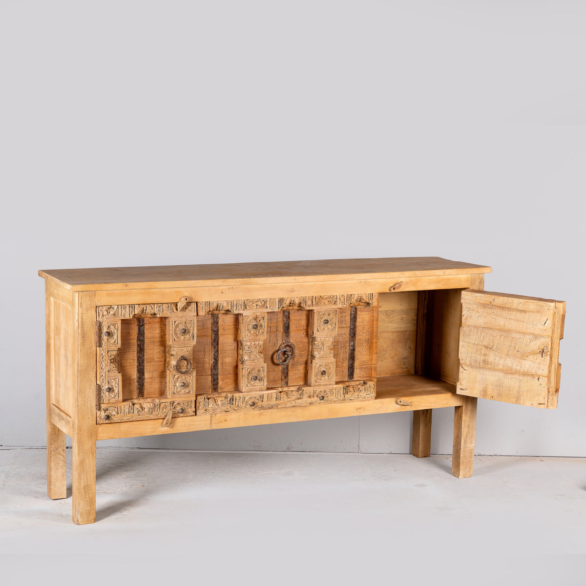 Side angle image with open door of a Kanan console table made from old teak door panels, featuring traditional hand-carved details, iron ring accents, and built-in storage in a natural rustic finish.