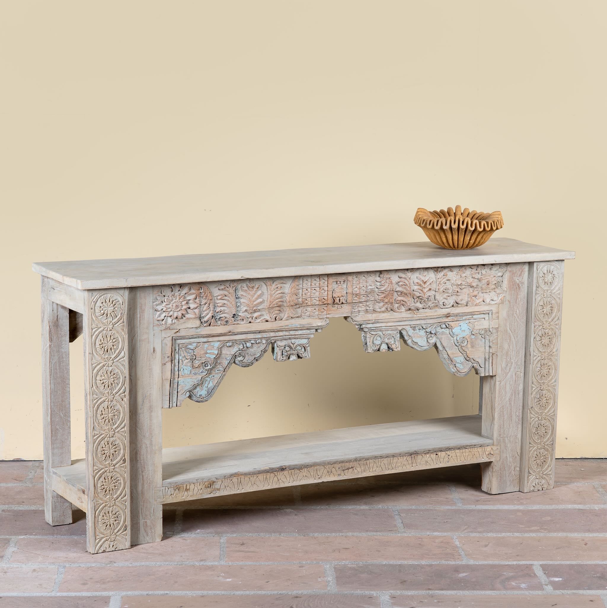 Side angle image of a Karam reclaimed teak console table with hand-carved Indian details, lower shelf, and vintage whitewashed finish.