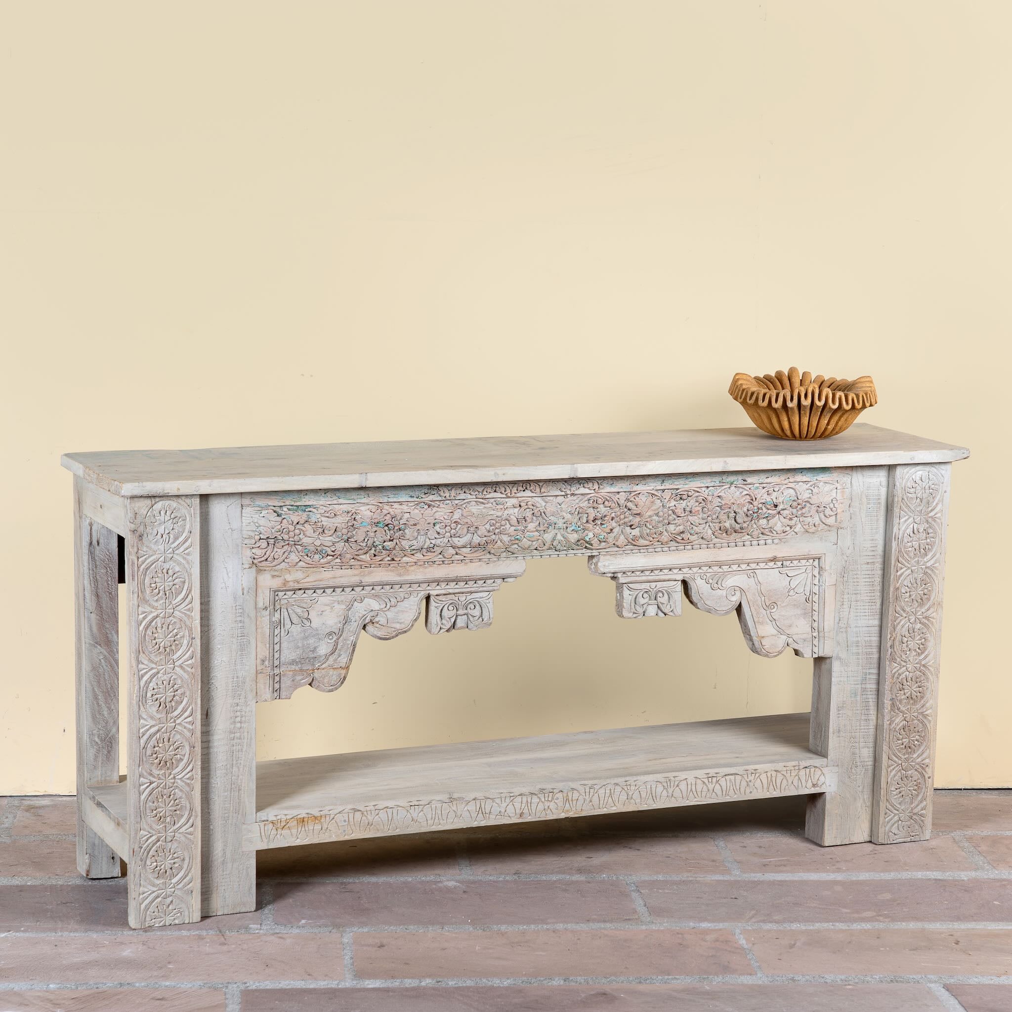 Side angle image of a Kayleb reclaimed teak console table with hand-carved Indian floral details, lower shelf, and whitewashed vintage finish.