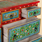 A close-up of a hand-painted wooden cabinet with open drawers, showcasing vibrant turquoise floral panels, a red upper frame, and black metal ring pulls.