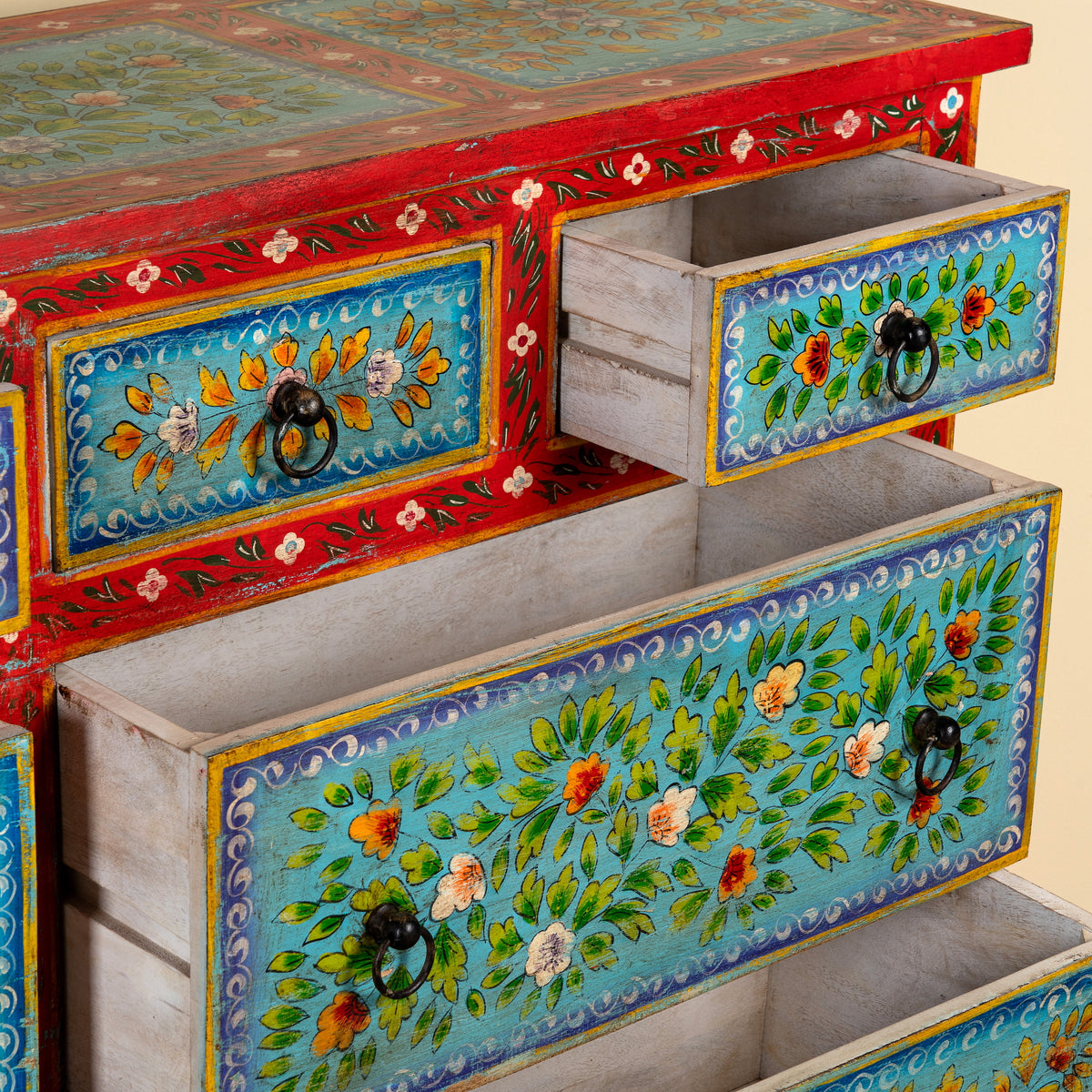 A close-up of a hand-painted wooden cabinet with open drawers, showcasing vibrant turquoise floral panels, a red upper frame, and black metal ring pulls.