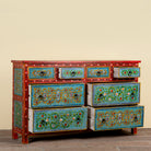 A hand-painted wooden dresser with six drawers, featuring a red frame and turquoise blue fronts decorated with intricate floral and geometric patterns, set against a plain beige wall.