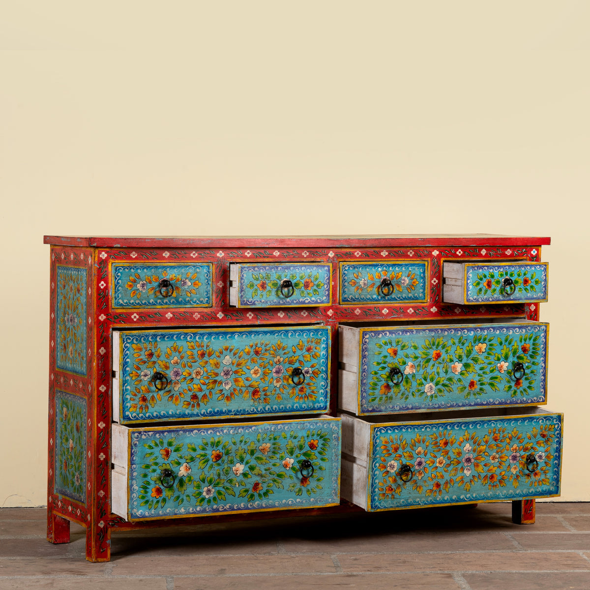 A hand-painted wooden dresser with six drawers, featuring a red frame and turquoise blue fronts decorated with intricate floral and geometric patterns, set against a plain beige wall.