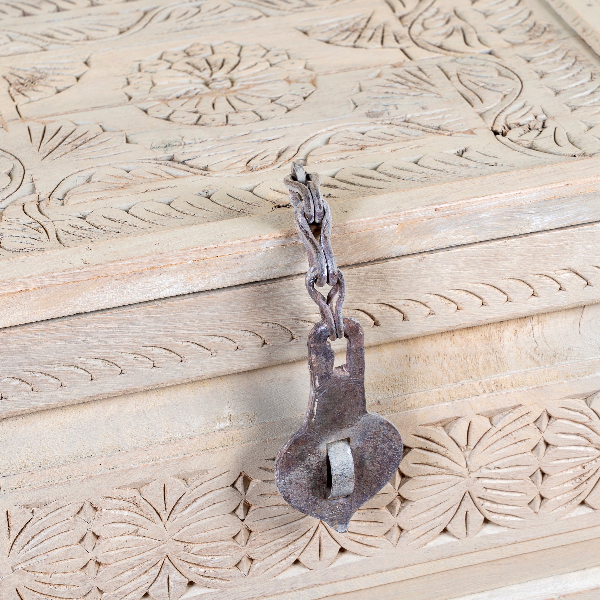 A rustic, dark metal padlock with a chain clasp resting on a light beige carved wooden surface.