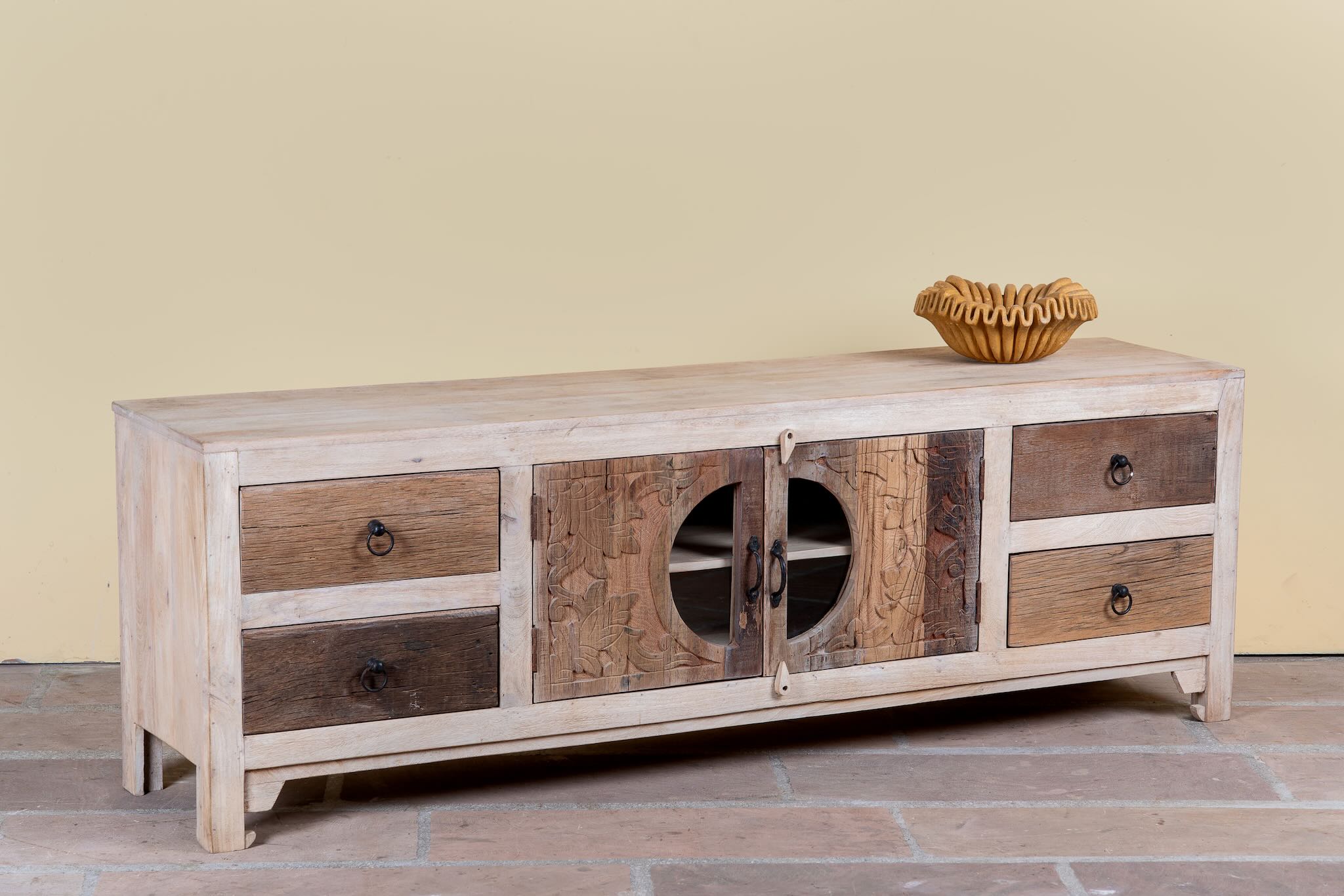 Side angle image of a Kishen handcrafted reclaimed wood lowboard with driftwood front detailing, carved cabinet doors, four drawers, and natural rustic boho style finish.