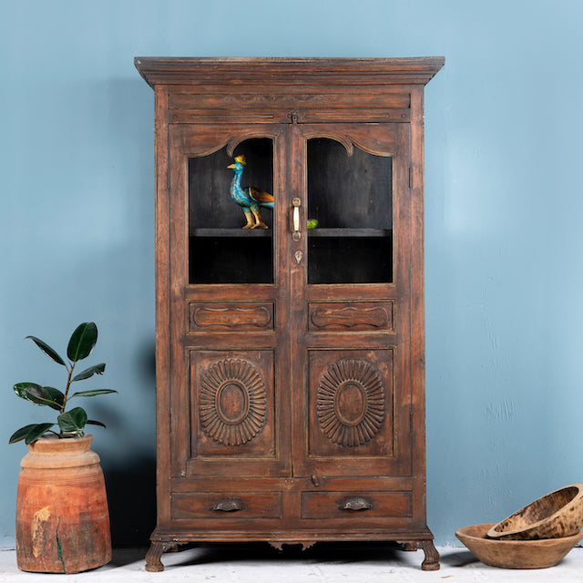 A tall, refurbished antique teak wood showcase with a glass front and a bird perched on a decorative element, placed against a light blue wall with a brown plant beside it.