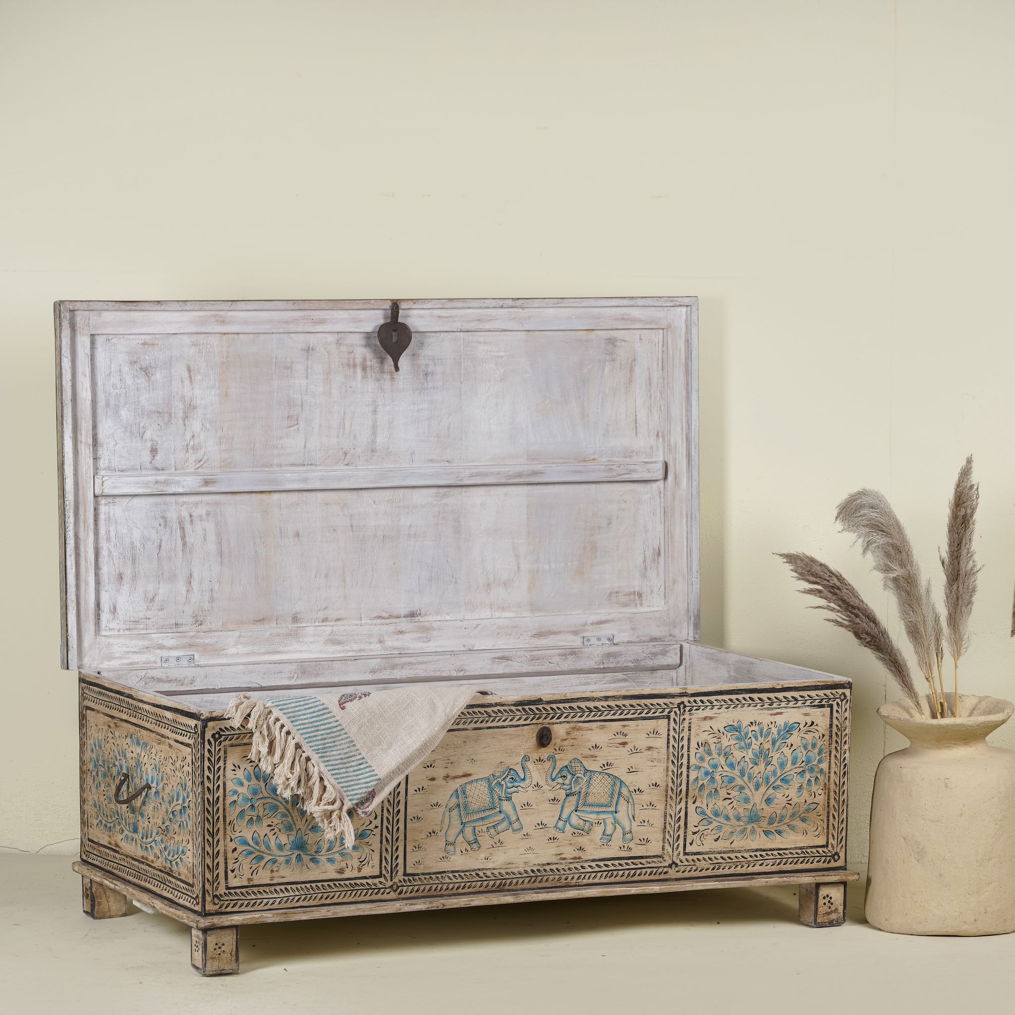 Image shows open lid of a large hand-painted chest made from old reclaimed wood, featuring intricate blue designs of elephants and floral motifs on a distressed beige background, with a central latch and a rustic vintage finish.
