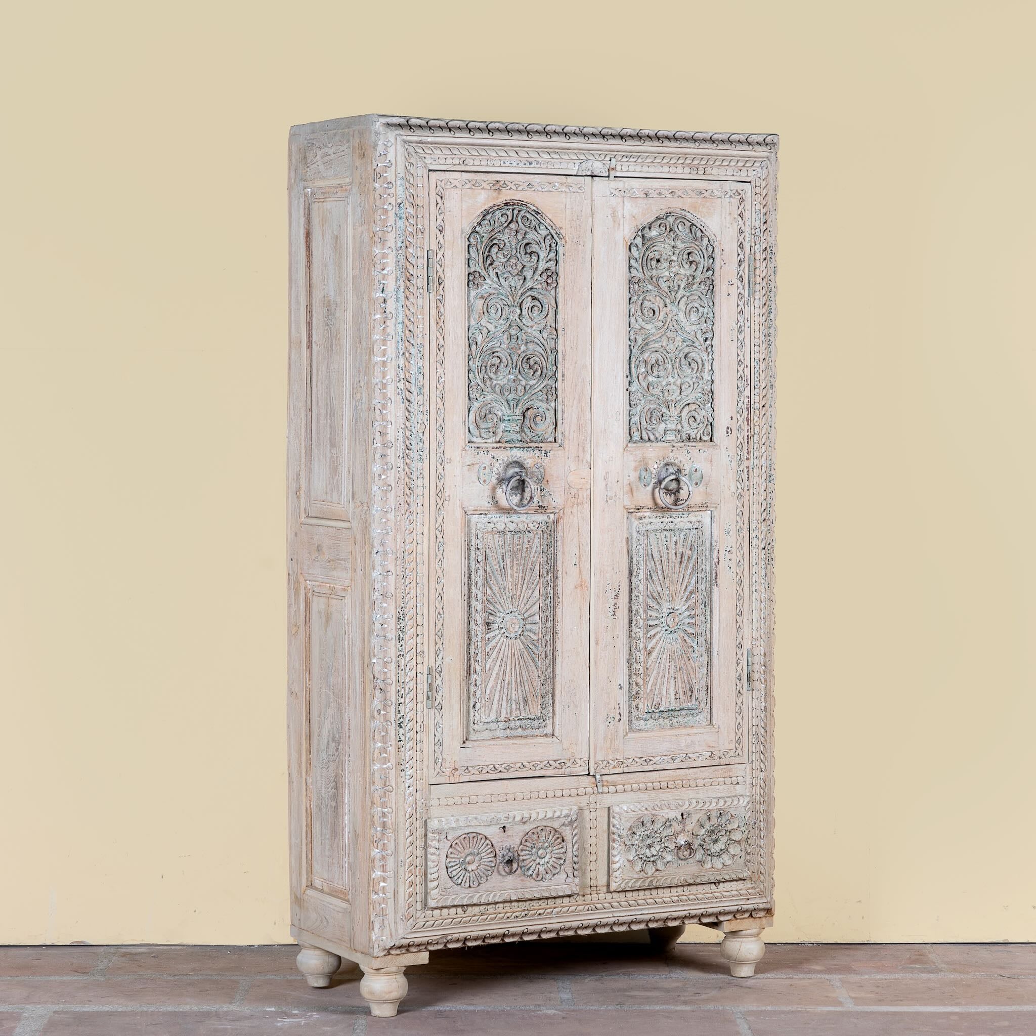 Side angle image of a Kyra antique reclaimed teak wood wardrobe with intricate Indian hand carvings, double doors, and vintage distressed finish.