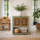 Lifestyle image of a small hand-painted console made from reclaimed wood, featuring vibrant decorative motifs in red, green, blue, and gold on double doors, with intricate floral and geometric patterns and a lower open shelf for added display.

