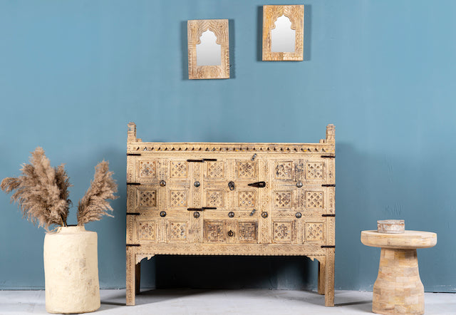 An antique Indian treasure chest in Ibiza style, made of teak wood with intricate hand-carved patterns and iron applications, displayed with decorative items and a vase in the background.