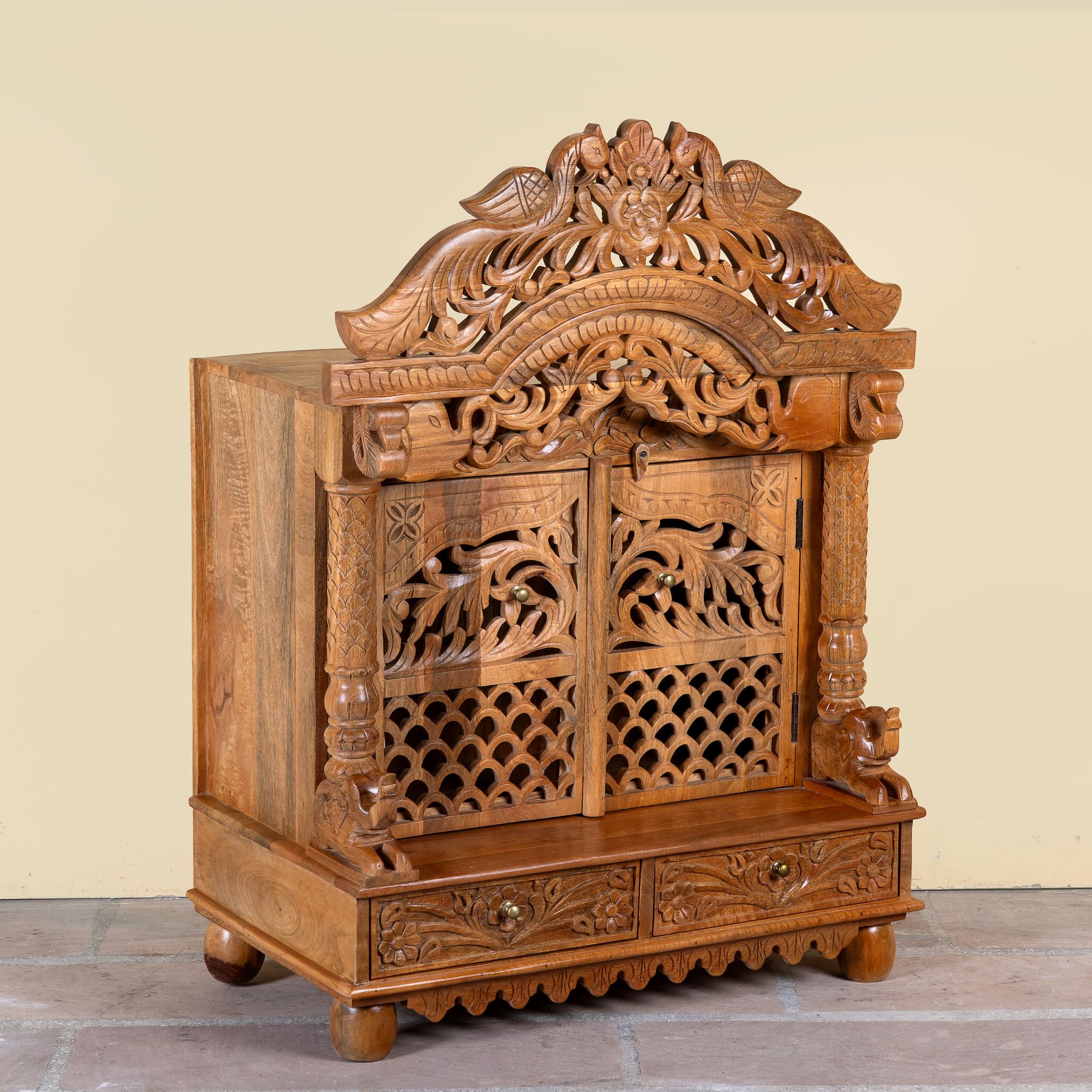 Side angle image of a hand carved mango wood Indian mandir temple with ornate floral carvings, lattice doors, and two storage drawers.